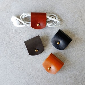 May include: Four leather cord organizers in different colors. The organizers are shaped like small squares with rounded corners and have a gold button in the center. The organizers are on a gray surface.