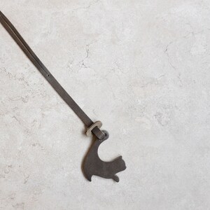 Peut inclure: Une sangle en cuir marron avec une boucle en m&eacute;tal et un charme en m&eacute;tal en forme de chat. La sangle est suspendue sur un fond gris clair.