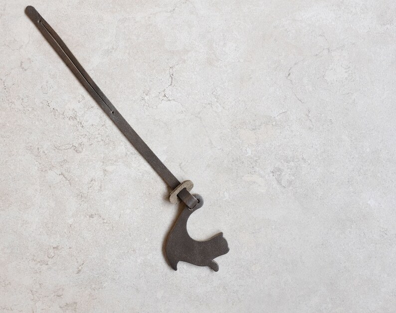 Peut inclure: Une sangle en cuir marron avec une boucle en m&eacute;tal et un charme en m&eacute;tal en forme de chat. La sangle est suspendue sur un fond gris clair.