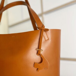 Peut inclure: Un sac cabas en cuir marron avec une bandouli&egrave;re en cuir et un petit charme en cuir en forme de poisson. Le sac a une couture cousue et le mot "clif" est emboss&eacute; sur le devant.