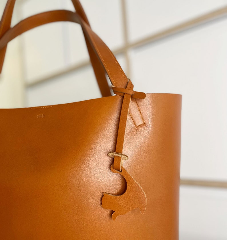 Peut inclure: Un sac cabas en cuir marron avec une bandouli&egrave;re en cuir et un petit charme en cuir en forme de poisson. Le sac a une couture cousue et le mot "clif" est emboss&eacute; sur le devant.