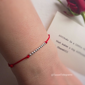 May include: A red string bracelet with silver beads. The bracelet is worn on a person's wrist. The text "@TasselTelegrams" is visible in the bottom right corner of the image.