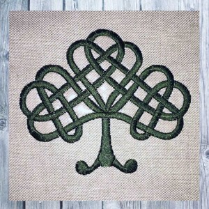 May include: A green Celtic knot tree design embroidered on a beige fabric. The tree features intricate knotwork in a symmetrical pattern, with a trunk and rounded base. The background is a textured, neutral-toned fabric.