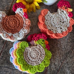 May include: Three sets of crocheted chicken-shaped coasters. One set features brown and cream yarn, another uses orange and cream, and the third displays a vibrant mix of green, yellow, blue, and pink. Each chicken has a red comb and orange beak.