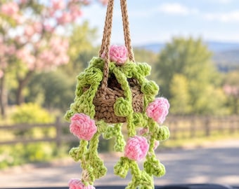 May include: A crocheted hanging plant ornament for a car rearview mirror, featuring a small brown pot, light green trailing vines, and several small pink flowers. The entire piece is suspended by two thin, braided brown yarn cords.