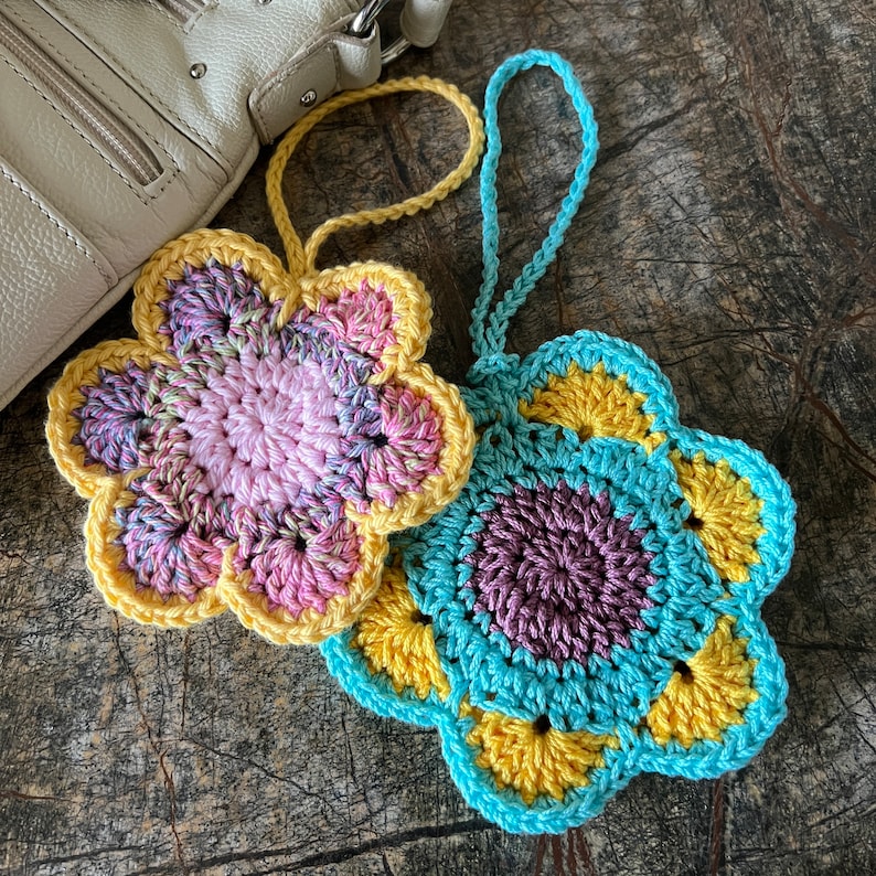 May include: Two handmade crochet flower ornaments with hanging loops. One flower features a light pink center with variegated pink and purple petals edged in yellow. The second flower has a dark purple center with yellow petals edged in light blue.