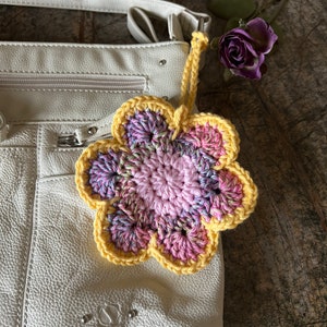 May include: A crocheted flower bag charm featuring a solid light pink center, variegated pink and purple petals, and a bright yellow border. The charm includes a matching yellow yarn loop for attachment and is displayed against a white leather bag.