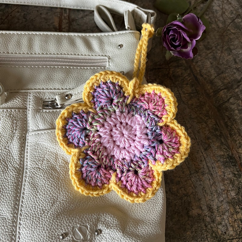 May include: A crocheted flower bag charm featuring a solid light pink center, variegated pink and purple petals, and a bright yellow border. The charm includes a matching yellow yarn loop for attachment and is displayed against a white leather bag.