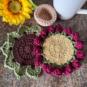 May include: Two round crochet coasters featuring floral designs. One coaster has a dark brown center with olive green petals, while the other has a pale yellow center with deep magenta petals. A small beige crochet bowl sits nearby.