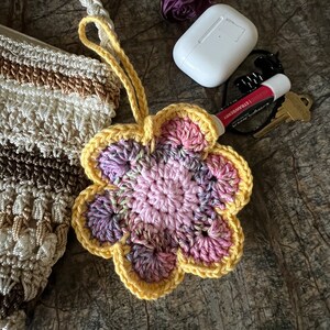 May include: A crocheted flower keychain with a yellow border and a variegated pink and purple center. The flower is attached to a loop and sits next to a white earbud case, a strawberry lip balm, a metal key, and a crocheted bag.