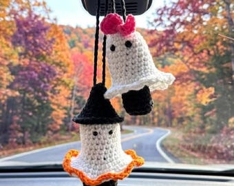 May include: Two crocheted ghost ornaments hanging from black strings. One ghost wears a black pointed hat and has an orange ruffled base. The other ghost features a pink bow on top. Both have white bodies and black bead eyes.