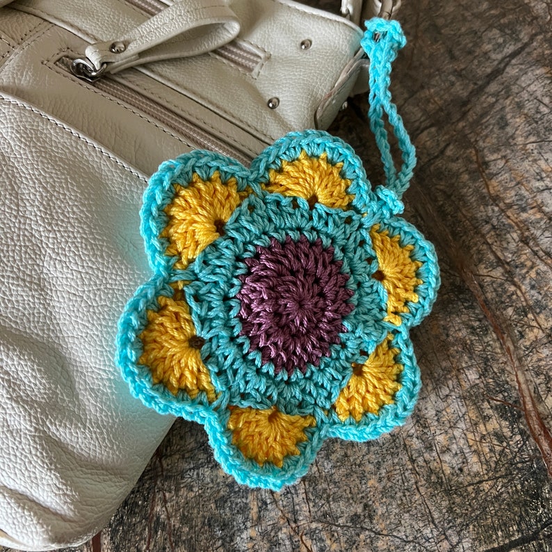May include: A crocheted flower ornament featuring a dark purple center, yellow petals, and a light blue border with a matching loop for hanging. The flower is constructed with yarn and displays a multi-layered, circular design with six distinct petal sections.