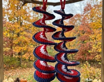 May include: Two hanging spiral ornaments crafted from red, white, and blue yarn. Each piece features a continuous crochet pattern with a central white core, a red middle section, and a dark blue outer edge, finished with a small loop at the top.