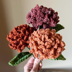 Puede incluir: Un ramo de tres flores de ganchillo en tonos naranja, &oacute;xido y burdeos. Las flores est&aacute;n unidas a tallos de ganchillo verdes.