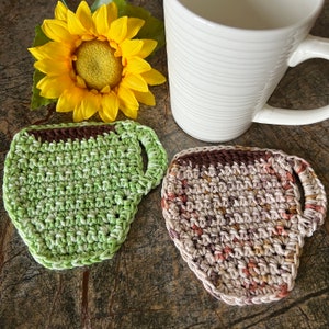May include: Two crocheted cup coasters in the shape of a coffee mug. One coaster is green and the other is brown with hints of orange and red.