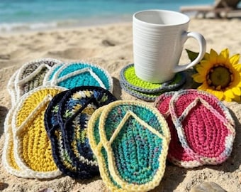 May include: A collection of crochet coasters shaped like flip-flops in various colors including yellow, teal, dark blue, and pink, alongside a white ceramic mug and a yellow sunflower on a sandy surface.