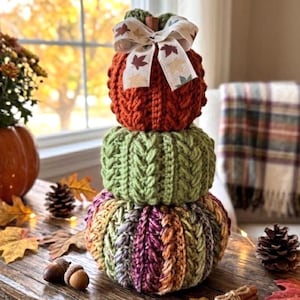 May include: A stacked pumpkin decoration made of three crocheted pumpkins in orange, green, and a variegated autumn-toned yarn. The top pumpkin is topped with a cream-colored ribbon featuring a brown and orange leaf pattern.