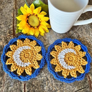 May include: Two handmade crochet coasters with a sunflower design. The coasters feature a blue border, yellow petals, and a white crescent moon shape in the center. A yellow sunflower and a white mug are also in the image.