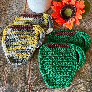 May include: Set of four crocheted cup coasters. Two coasters are green and two are gray, yellow, and white. The coasters are shaped like coffee mugs.