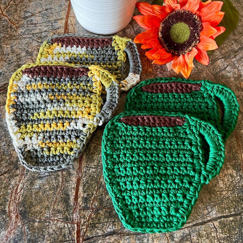 May include: Set of four crocheted cup coasters. Two coasters are green and two are gray, yellow, and white. The coasters are shaped like coffee mugs.