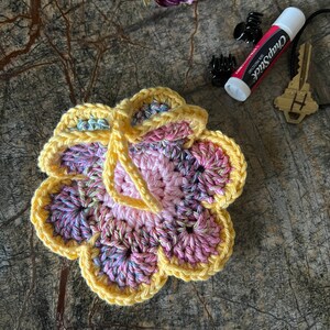 May include: A crocheted flower ornament featuring a light pink center surrounded by variegated pink, purple, and blue petals, all edged in bright yellow yarn. A yellow yarn loop is attached to the top for hanging purposes.
