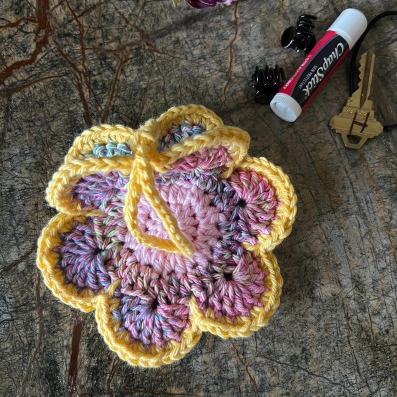 May include: A crocheted flower ornament featuring a light pink center surrounded by variegated pink, purple, and blue petals, all edged in bright yellow yarn. A yellow yarn loop is attached to the top for hanging purposes.