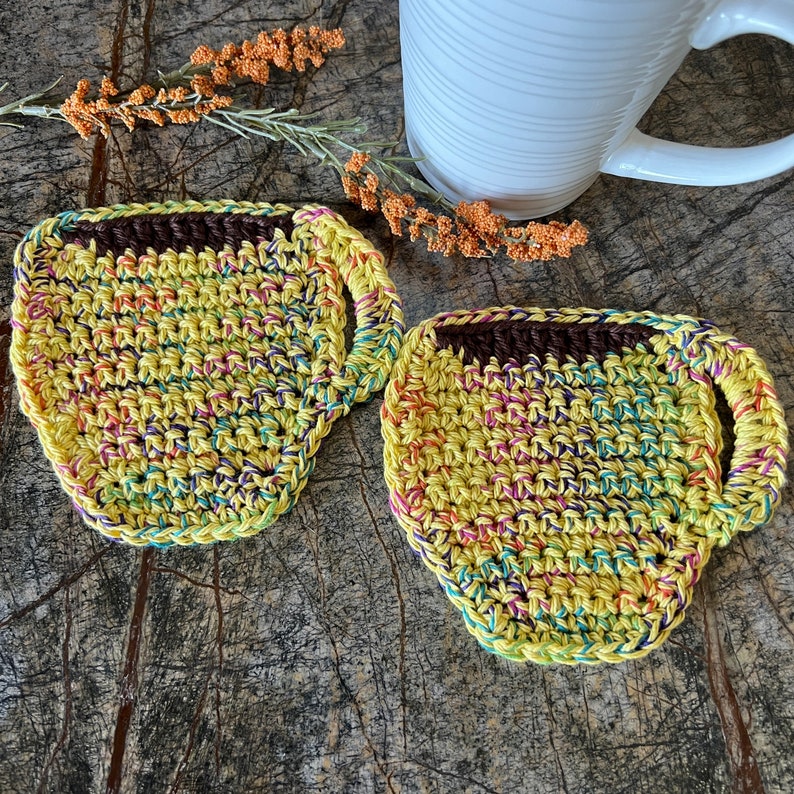 May include: Two crocheted cup coasters in a yellow, green, and purple color scheme. The coasters are shaped like coffee cups and have a brown crocheted rim.