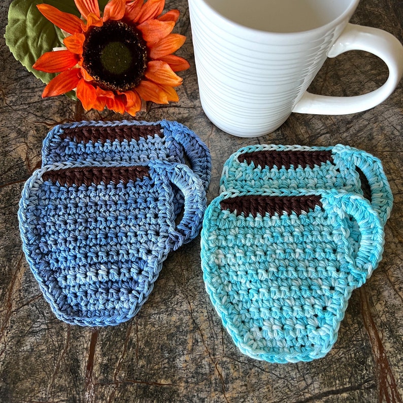 May include: Two sets of crocheted cup coasters in blue and turquoise shades. Each set has two coasters shaped like coffee mugs with brown crocheted handles.