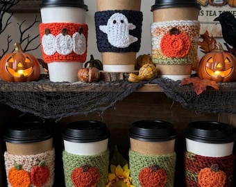 May include: A collection of eight disposable coffee cups featuring various handmade crochet sleeves. The sleeves display seasonal motifs including white ghosts, orange pumpkins, and autumn-colored yarn patterns in shades of green, brown, and cream.