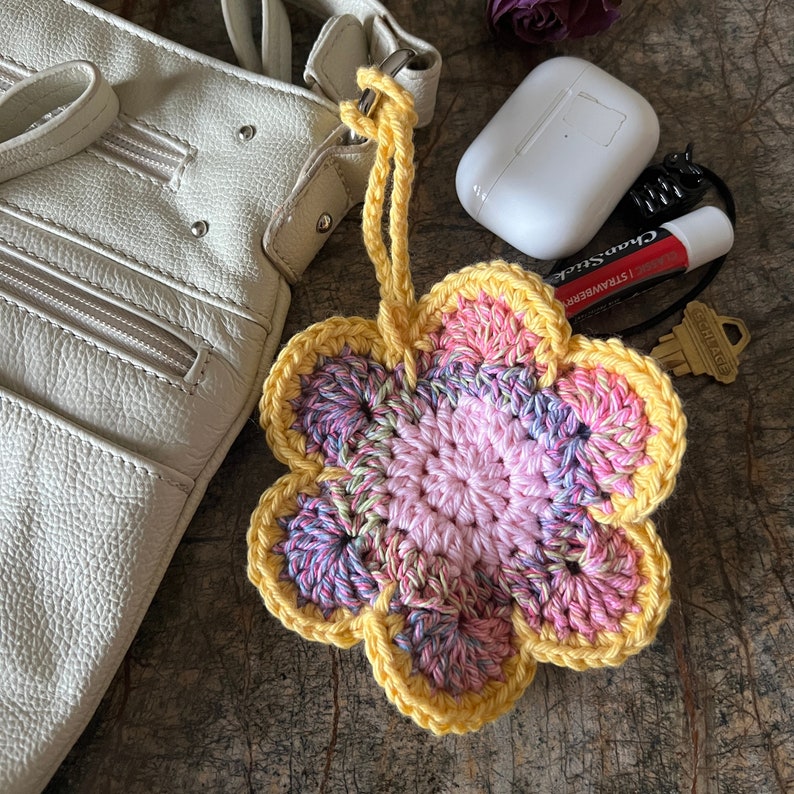 May include: A crocheted flower keychain featuring a central pink section surrounded by variegated pink, purple, and green petals, all edged in bright yellow yarn. The flower is attached to a yellow yarn loop and a metal clasp.