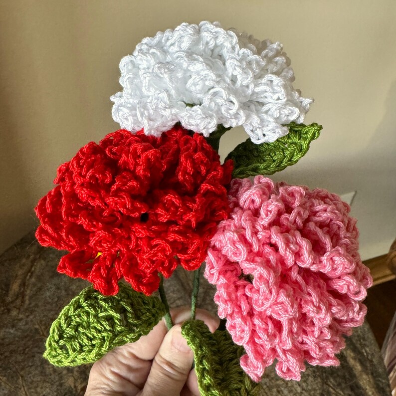 Puede incluir: Un ramo de tres flores de ganchillo en rojo, blanco y rosa. Las flores est&aacute;n hechas con un punto texturizado y rizado y tienen hojas verdes de ganchillo.