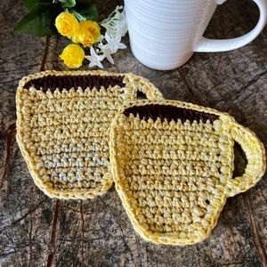 May include: Two crocheted cup coasters in a yellow and brown color scheme. The coasters are shaped like coffee cups with a handle.