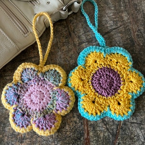 May include: Two handmade crochet flower ornaments with hanging loops. The left flower features a pink center with variegated pink, blue, and yellow petals edged in yellow. The right flower has a purple center with yellow petals edged in light blue.