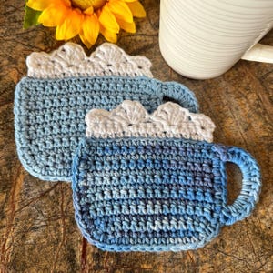 May include: Two crocheted blue and white mug cozies with a scalloped edge. The cozies are made with a textured stitch and have a handle.