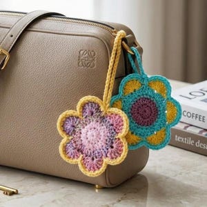 May include: A beige leather handbag features two hanging crochet flower charms. One charm has a pink and purple center with a yellow border, while the other has a purple center with yellow and teal petals. Both are attached by yellow crochet loops.