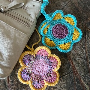 May include: Two handmade crochet flower ornaments with hanging loops. One flower features a purple center with yellow and light blue petals. The second flower has a pink center with variegated pink and yellow petals.
