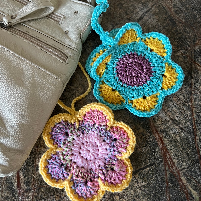 May include: Two handmade crochet flower ornaments with hanging loops. One flower features a purple center with yellow and light blue petals. The second flower has a pink center with variegated pink and yellow petals.