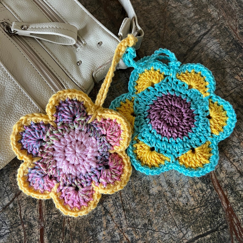 May include: Two crocheted flower-shaped ornaments. One flower features a pink center with variegated pink and purple petals edged in yellow. The second flower has a dark purple center with turquoise petals and yellow accents.