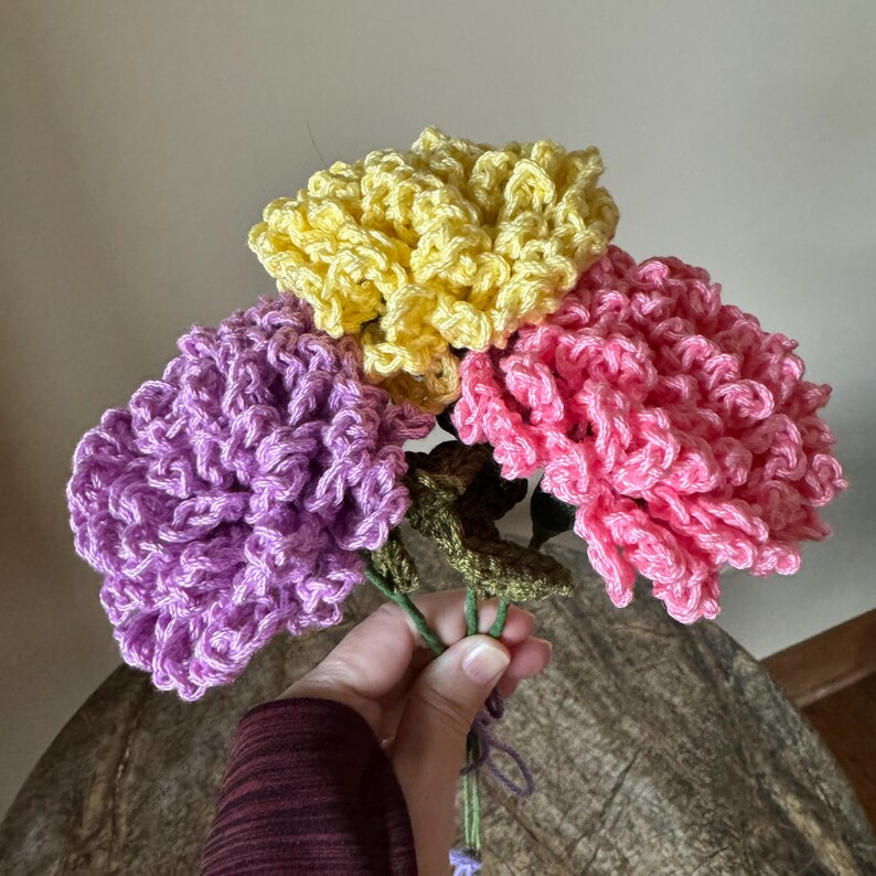 Puede incluir: Un ramo de tres claveles de ganchillo en tonos de morado, rosa y amarillo. Los claveles est&aacute;n atados con hilo verde.