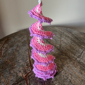 May include: A crocheted spiral made of pink and white yarn. The spiral is made of multiple layers of crocheted fabric, each layer slightly smaller than the one below it. The spiral is standing upright on a wooden surface.