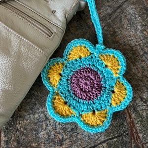 May include: A crocheted flower ornament featuring a central purple circle, surrounded by yellow petals, and outlined in light blue yarn. The flower includes a matching light blue crocheted loop for hanging, attached to a light beige textured bag.