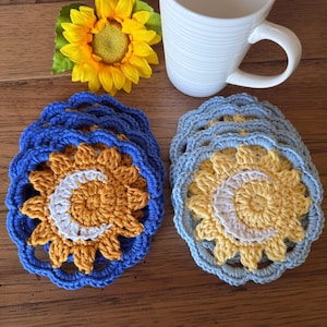 May include: A set of round, crocheted coasters featuring a central sun and crescent moon design. One stack has dark blue borders with gold centers, and the other has light blue borders with yellow centers. A white mug and a sunflower are nearby.