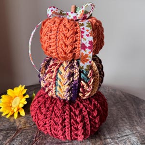 May include: A stack of three crocheted pumpkins in shades of orange, burgundy, and multicolored yarn. The top pumpkin is orange, the middle one is a mix of colors, and the bottom is burgundy. A ribbon with fall leaves accents the top pumpkin.