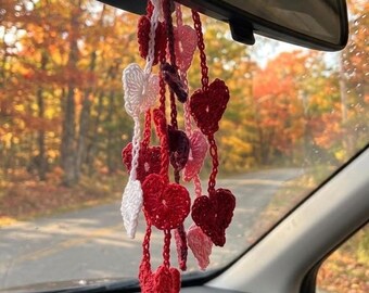 May include: A hanging car accessory featuring multiple crocheted hearts in shades of red, pink, white, and dark maroon. The hearts are attached to thin, crocheted strings of varying lengths, creating a cascading, layered arrangement.