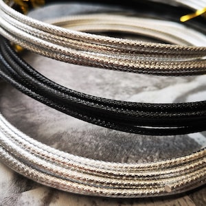 Könnte beinhalten: Nahaufnahme von mehreren aufgerollten Schnüren in Silber und Schwarz. Die Schnüre haben ein strukturiertes, gewebtes Aussehen und sind in sich überlappenden Kreisen angeordnet. Die Enden der Schnüre sind mit kleinen goldfarbenen Verschlüssen gesichert.