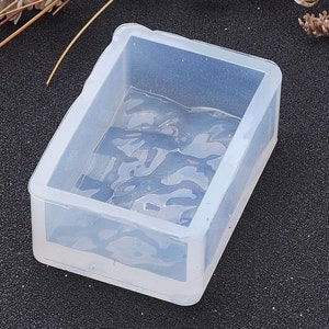 May include: A clear, rectangular silicone mould with a camouflage pattern inside. The mould has a box shape with slightly rounded corners. The background is a dark, speckled surface with dried flowers.