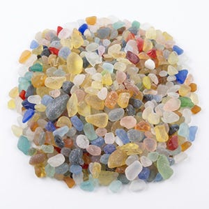 May include: A large collection of small, smooth, tumbled sea glass pieces in a variety of colors including yellow, blue, red, green, white, and clear. The pieces are rounded and frosted, arranged in a loose, circular pile on a white background.