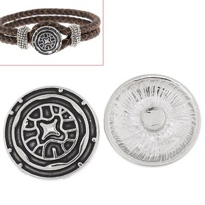 May include: A brown braided leather bracelet with a silver-toned circular clasp featuring a black recessed geometric pattern and two beaded silver-toned accents. Also shown are two views of the circular silver-toned button with a black center.