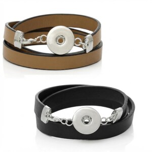 Könnte beinhalten: Zwei Wickelarmbänder, eines in Hellbraun und eines in Schwarz, jeweils mit einem glatten, runden Druckknopf aus silberfarbenem Metall in der Mitte, der durch kleine silberfarbene Metallketten und rechteckige Endkappen mit dem Band verbunden ist.