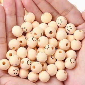 May include: A collection of small, light-coloured wooden beads with drilled holes. Each bead features a hand-painted smiley face with black eyes and a curved mouth. The beads are held in a person's hand.